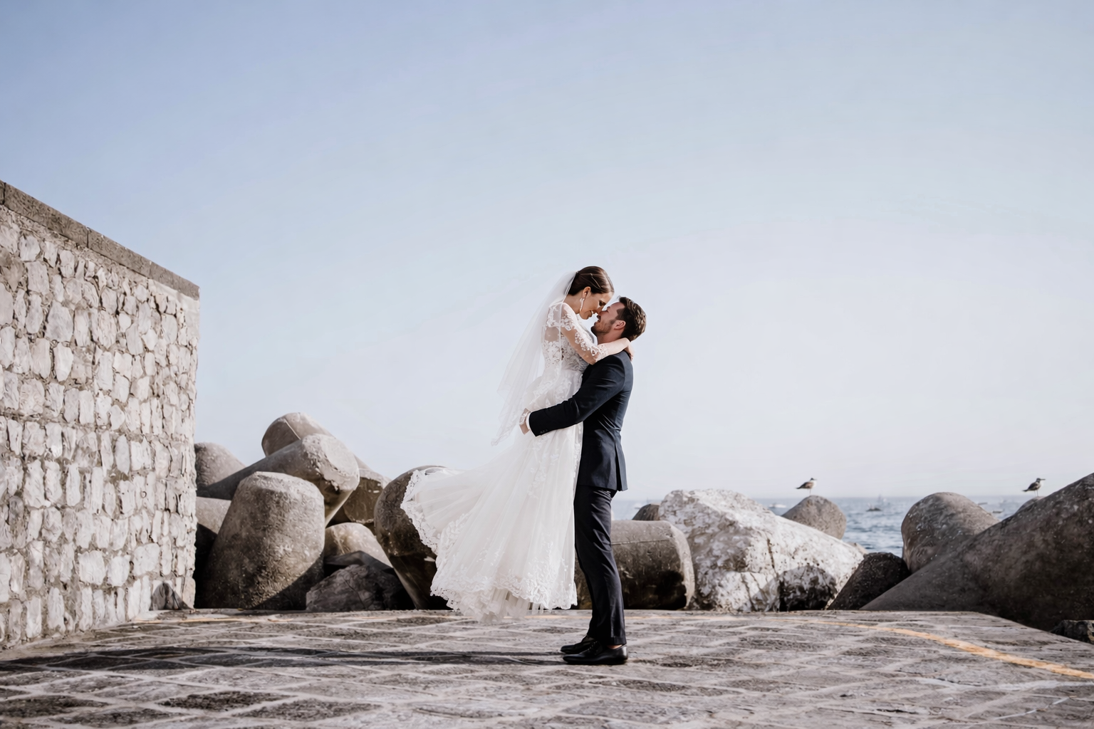 groom lifting bride on seaside pier with rocks and sea in Amalfi Coast minimal background